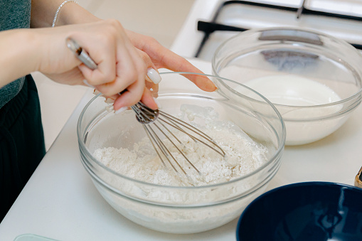 The hand of one young Caucasian unrecognizable girl manually mixes the dough with a whisk in a glass bowl, standing at a white table, close-up side view. Step-by-step instructions for baking synabons. Step 2.