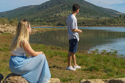 A couple, she sitting and he standing, contemplates the beauty of the landscape, a lake surrounded by beautiful forest, a vacation spot where they can spend a honeymoon