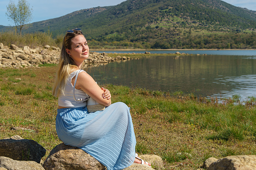 Portrait of a beautiful elegant woman sitting on the shore of Lake La Pinilla, a relaxing landscape for a summer vacation