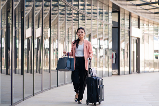Young Asian female traveler carrying shopping bags while pulling wheeled suitcases at airport terminal. The business woman shopping at airport duty free outlets upon her arrival.