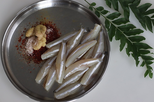 Freshly cut and cleaned anchovy fish presented on a steel plate along with chili powder, pepper, ginger garlic paste and salt for marination, surrounded with fresh curry leaves.