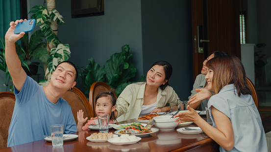 Young Asian family using mobile phone taking selfie and having fun sitting at dining table at backyard outside home. Multi-generation family enjoying spending together concept.
