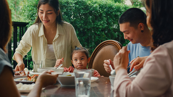 Asian family eating chinese food and having fun sitting at dining table at backyard outside home. Multi-generation family enjoying spending together concept.