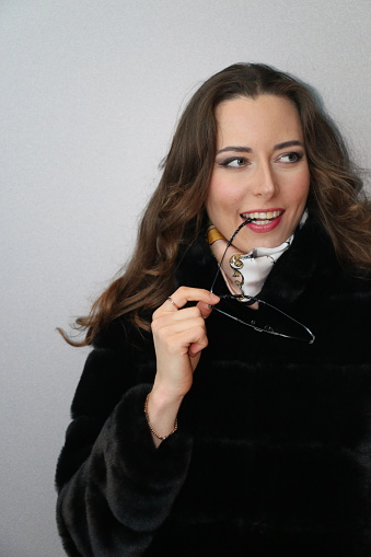 A Ukrainian woman looking to the side with a smile thinking about something. She is wearing long brown hair, makeup, holding sunglasses, has a scarf and black coat.