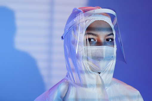 Medium close up shot female laboratory technician dressed in hazmat suit, face shield and mask looking at camera while standing at blue background with blinds shadow