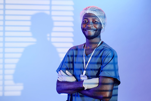Medium shot of cheerful Black man surgeon in medical mop cap standing hands crossed in pastel blue light and blinds shadow while looking at camera