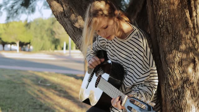 A blonde from Europe plays the guitar. Fingering of complex notes. A schoolgirl sits on a tree in the park. Musical education. The child is learning music
