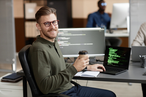 Medium long shot of smiling Caucasian male programmer in glasses looking at camera and holding takeaway coffee cup while sitting at desk in cybersecurity department