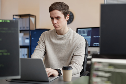 Medium shot of busy Caucasian male IT specialist in pastel sweater working on laptop while sitting at desk in cybersecurity office