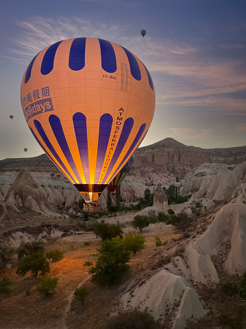 a hot air balloon flies at ground level between the fairy chimney at dawn in Cappadocia, Turkey, the glow of the burner illuminates the balloon and the ground, vertical