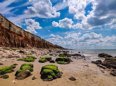 Scenic beach view of white and orange chalk and limestone cliffs at Old Hunstanton, Norfolk, England. Sand, sea, blue sky, white clouds and green rocks covered in seaweed