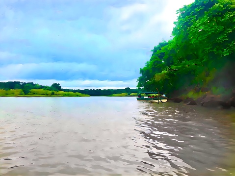 A boat in the Panama Canal next to the jungle with tall trees and bright green leaves. The. Water is muddy brown and has small ripples. There is a grassland with more trees in the background. Sky is blue with clouds.