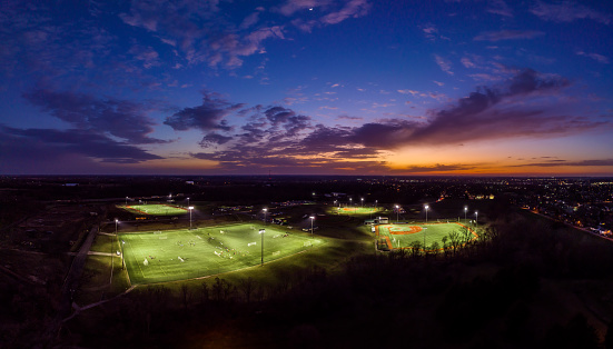 An aerial panoramic photo via drone with a colorful twilight sky at dusk over the sports complex with soccer, football, baseball, and softball fields lit up for play in the dark.