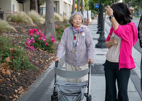 Senior woman walking using a mobility walker on the pedestrian footpath, chatting to her daughter. California.