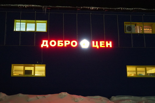 Petrozavodsk, Karelia, Russia, January 11, 2024: building - hangar of the Dobrotsen store, at night in winter. Red-yellow glow-in-the-dark letters and words on the facade. Saving money. Low prices.