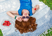 Portrait of a beautiful stylish woman with gorgeous hair lying on a picnic blanket with a red strawberry in her hand