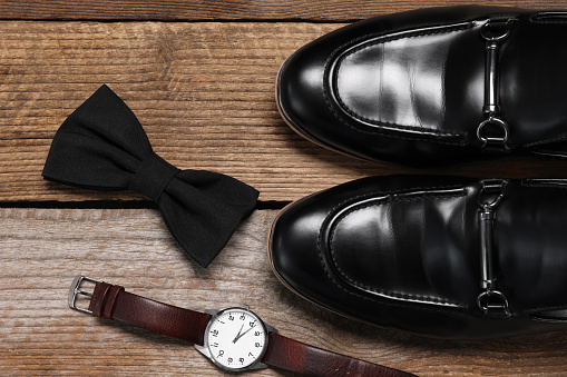 Stylish black bow tie, wristwatch and shoes on wooden background, flat lay