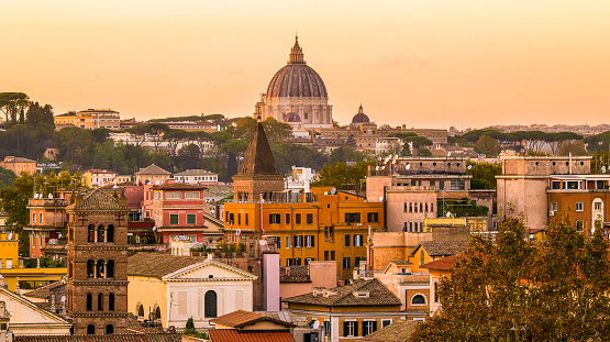 A splendid cityscape at sunset of the historic center of Rome seen from the terrace of the Aventine Hill gardens, in the heart of the Eternal City. The majestic dome of St. Peter's Basilica stands out on the horizon, while in the foreground the bell towers and buildings of the ancient Trastevere district. The Aventine Hill, one of the seven ancient hills of Rome, is one of the most visited places in the city due to the presence of gardens, Roman archaeological sites and some of the Byzantine and medieval churches, among the oldest in Rome. In 1980 the historic center of Rome was declared a World Heritage Site by Unesco. Image in original 16:9 ratio and high definition format.