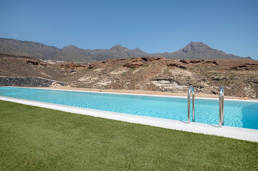 Luxurious long swimming pool overlooking the arid volcanic mountains, surrounded by a perfectly trimmed lawn, tranquil atmosphere of a high-end residential property suited for a lavishing lifestyle.