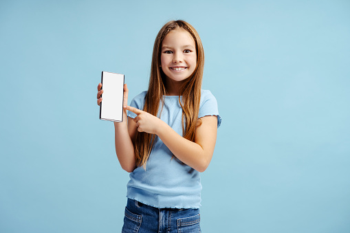 Portrait of happy little girl wearing casual clothes holding mobile phone and pointing at blank screen standing isolated on blue background. Technology concept