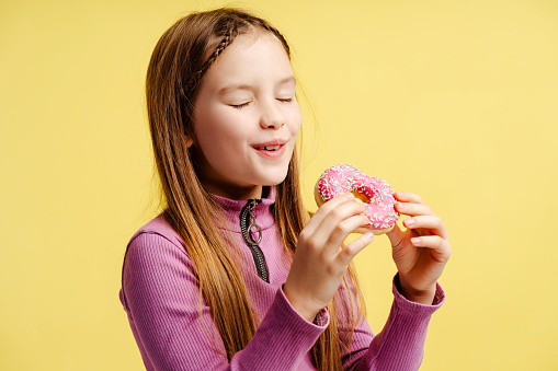 Satisfied beautiful girl holding pink donut, eating, feeling happy face isolated on yellow background. Tasty food concept