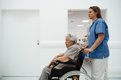 Portrait of nurse pushing senior patient in a wheelchair across hospital corridor, hall. Emotional support for elderly woman.