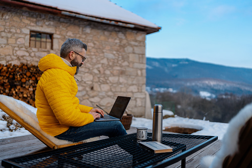 Man working from cozy cabin in mountains, sitting on terrace with laptop, enjoying cup of coffee. Concept of remote work from beautiful, peaceful location. Hygge at work.