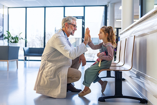Friendly pediatrician giving high five to little patient. Cute preschool girl in greeting doctor in hospital corridor.Concept of children healthcare and emotional support for child patients.