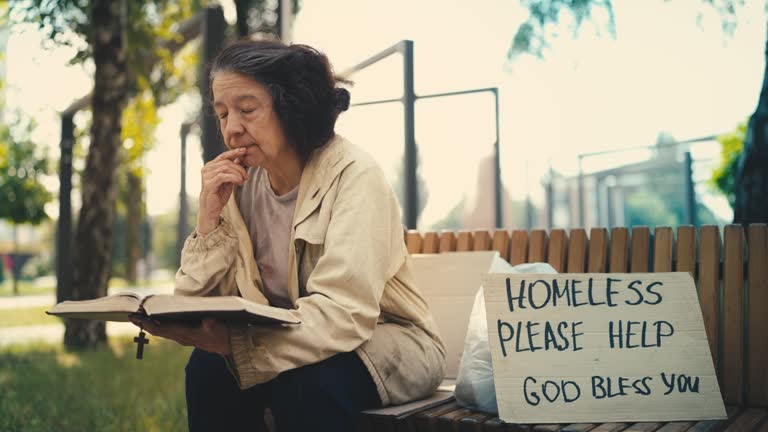 Religious homeless old woman reading a bible on bench, please help sign nearby