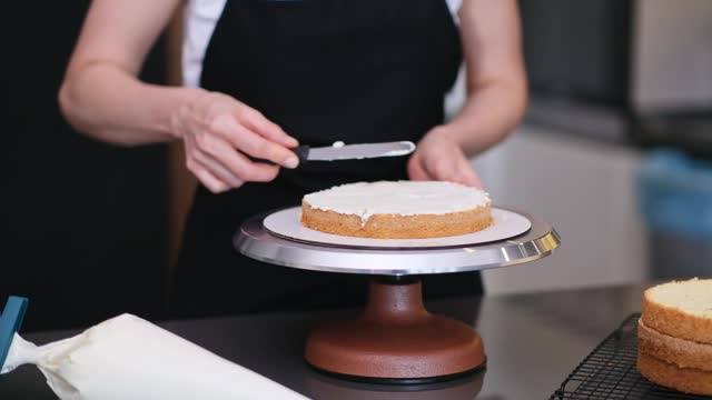Skilled female confectioner smearing organic cake layers with knife standing at table
