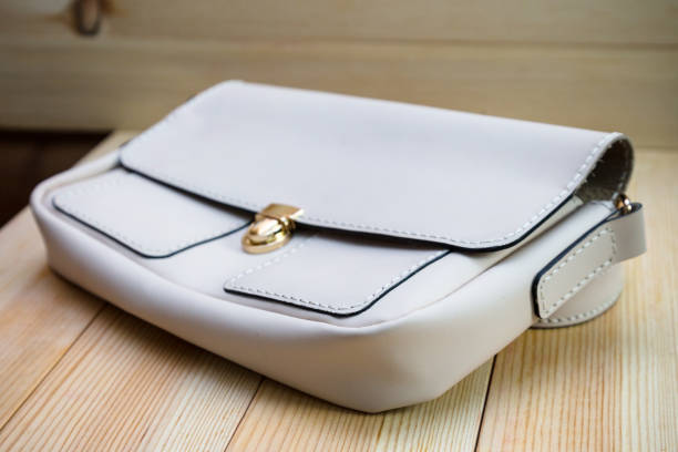 White leather women's handbag with a strap on a light wood background. An elegant accessory for stylish women. Handbag made genuine leather, comfortable and practical.