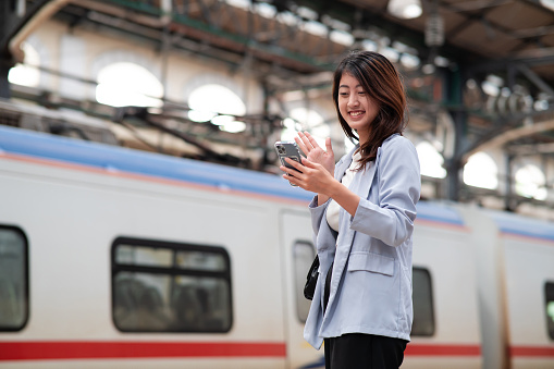 Young female Asian business traveler video calling using smart phone while waiting for train on railway station in the morning.