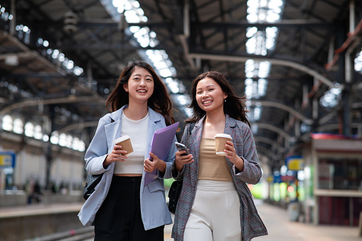 Two female Asian business travelers enjoy hot coffee while walking on the railway station platform. The two business women waiting for the train to go for work at Kuala Lumpur Railway Station in the morning.