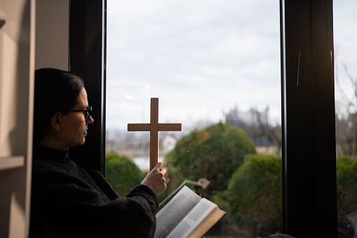 Woman holding wooden religious cross while sitting by the window and reading Holly Bible at home