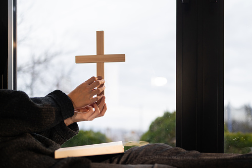 Close-up shot of unrecognizable woman holding religious cross against the window and over Holly Bible at home