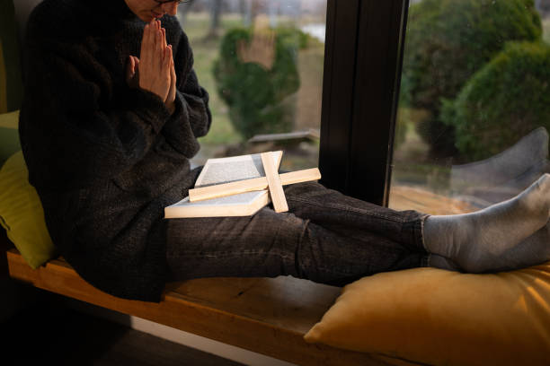 Woman sitting by the window and praying while holding Bible and wooden cross on her lap at home