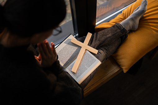 Woman sitting on window sill and praying over Holly Bible and wooden religious cross at home