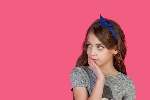 Portrait of Puzzled little cover girl with hand on cheek at pink isolated, confused looking away. Surprised kid girl lady model 9 year old, studio shot. Child shock emotion concept. Copy ad text space