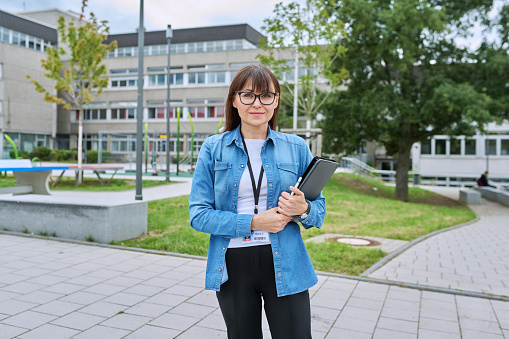 Middle-aged confident woman school teacher, mentor, pedagogue, psychologist, counselor, social worker with digital tablet in hands posing near school building, outdoor. Education, teaching, school
