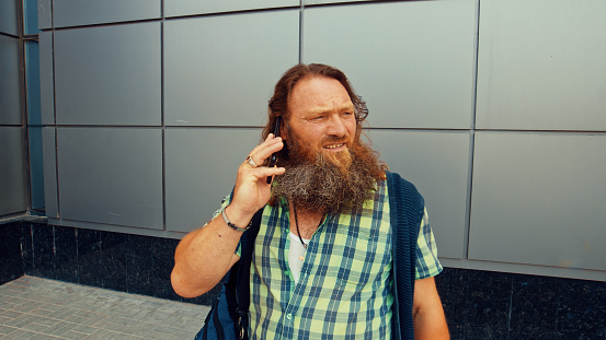 Red-bearded man talking on the phone standing near the gray wall