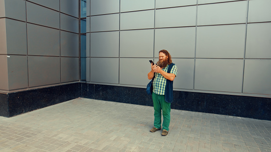 Elderly long-bearded man standing against a wall reading a message on a smartphone