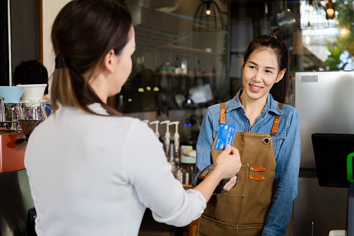 Asian waitress barista worker accepts credit card cashless payments from customer female in cafe restaurant, happy woman open bakery coffee shop, small business entrepreneur start-up lifestyle
