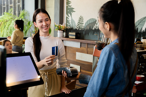 Asian waitress barista worker accepts credit card cashless payments from customer female in cafe restaurant, happy woman open bakery coffee shop, small business entrepreneur start-up lifestyle