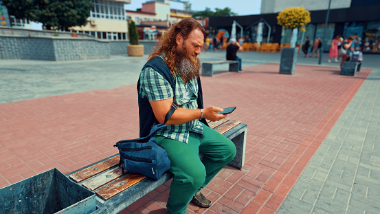 Redhead bearded man with long hair reading message on a phone sitting on a city bench