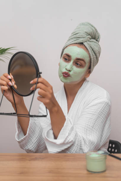 woman with green clay mask on face and towel wrap on head sending kiss to mirror reflection - mirror women kissing human face - fotografias e filmes do acervo