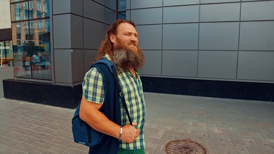 Senior redhead man with a long beard and hair walks through the city along a gray building wall