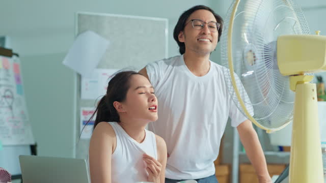 Young woman and friend enjoying air flow from fan at workplace Business person suffering from heat in front of small fan at workplace asian officer happy with electric fan standing at workplace