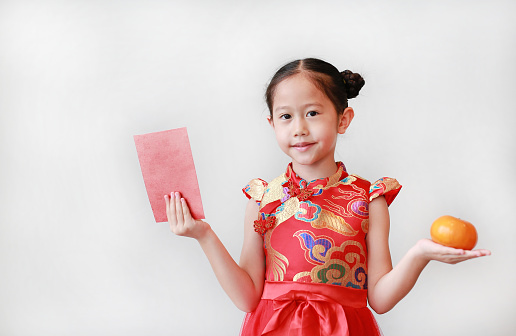 Beautiful asian little girl wearing traditional red cheongsam holding a red red envelope and orange fruit in hands isolated on white background. Chinese New Year celebration concept.