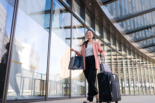 Young Asian female traveler carrying shopping bags while pulling wheeled suitcases at airport terminal. The business woman shopping at airport duty free outlets upon her arrival.