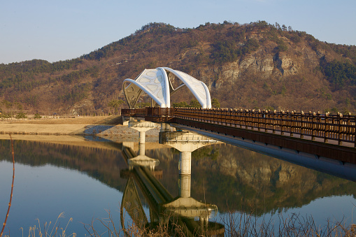 Sangju City, South Korea - March 9, 2017: The elegant Gyeongcheon Bridge, with its distinctive white metal awnings, spans gracefully over the mirror-like Nakdong River, leading to Gyeongcheon Island Park with the rugged Bibong Mountain in the backdrop.